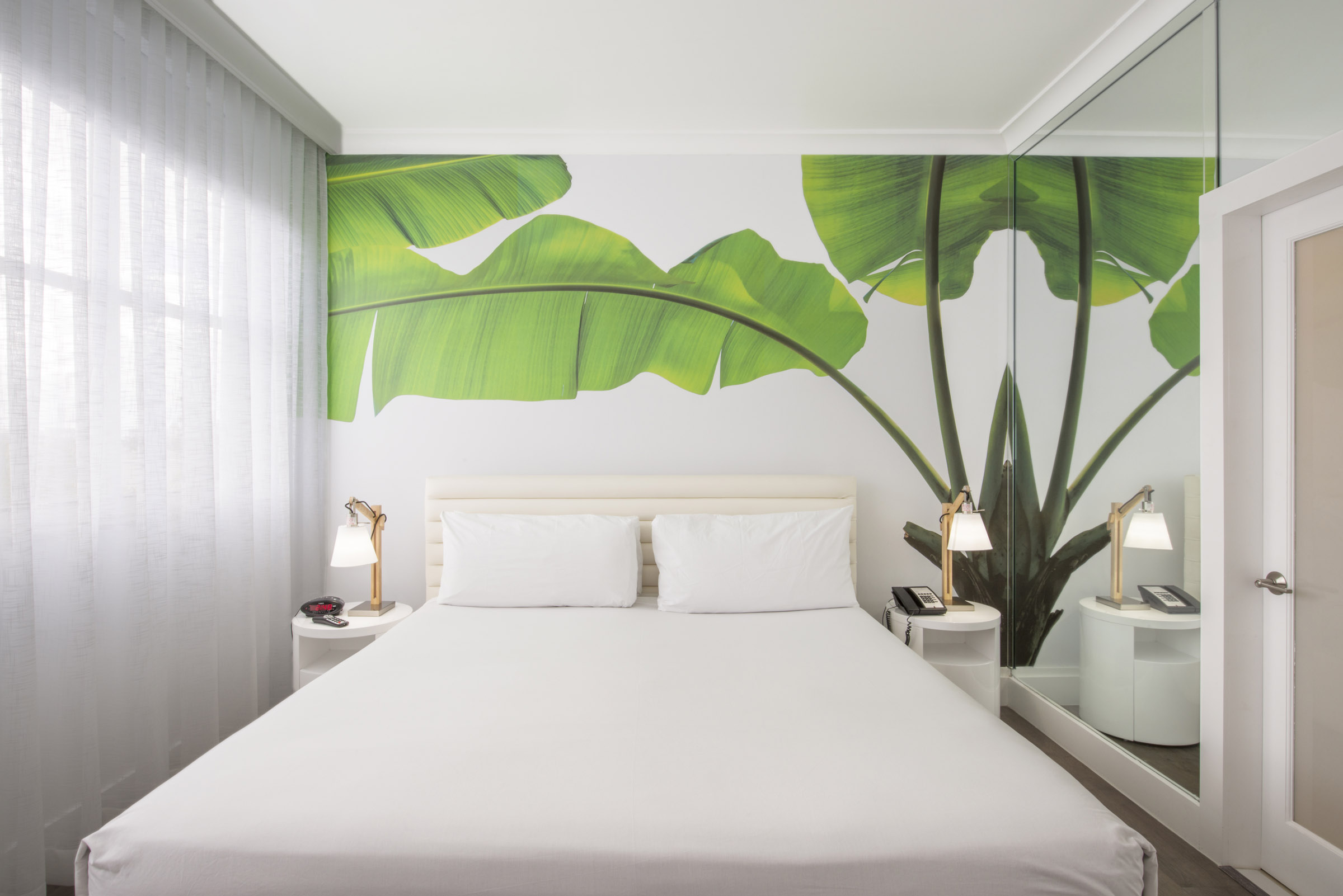 A neatly made bed with white linens and pillows in a room with a large window, tropical wallpaper, and two nightstands with lamps.