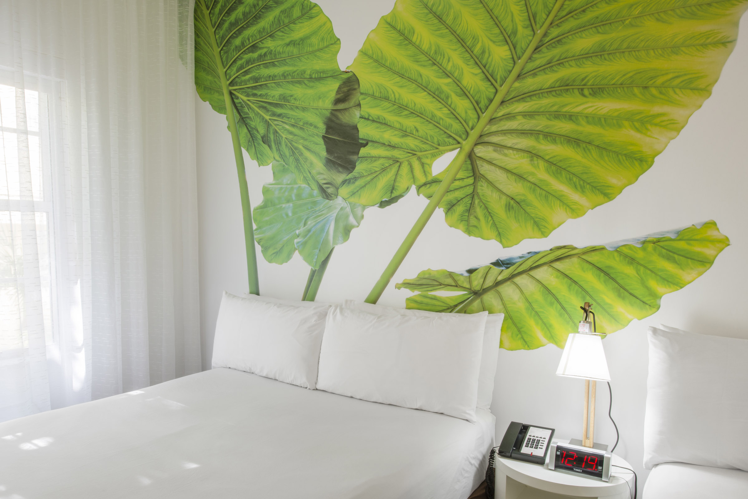A neatly made bed with white linens and pillows, a nightstand with a lamp, phone, and clock, in a room with a large leaf mural on the wall.
