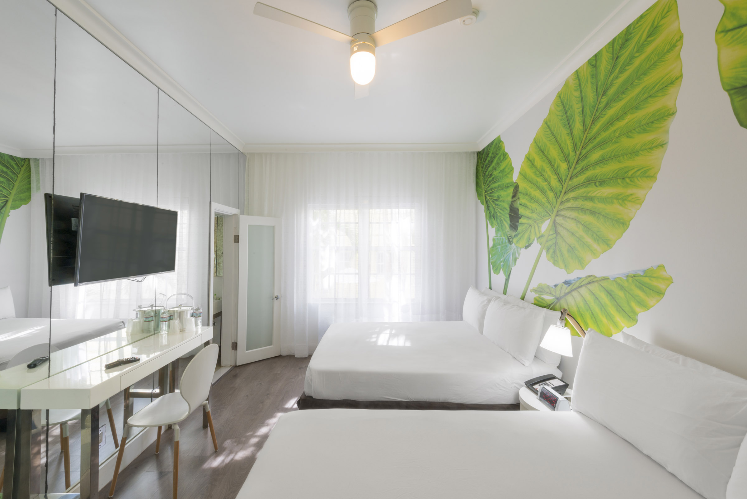 A bright and airy hotel room with two double beds, a desk, and a large leaf decorative wall.