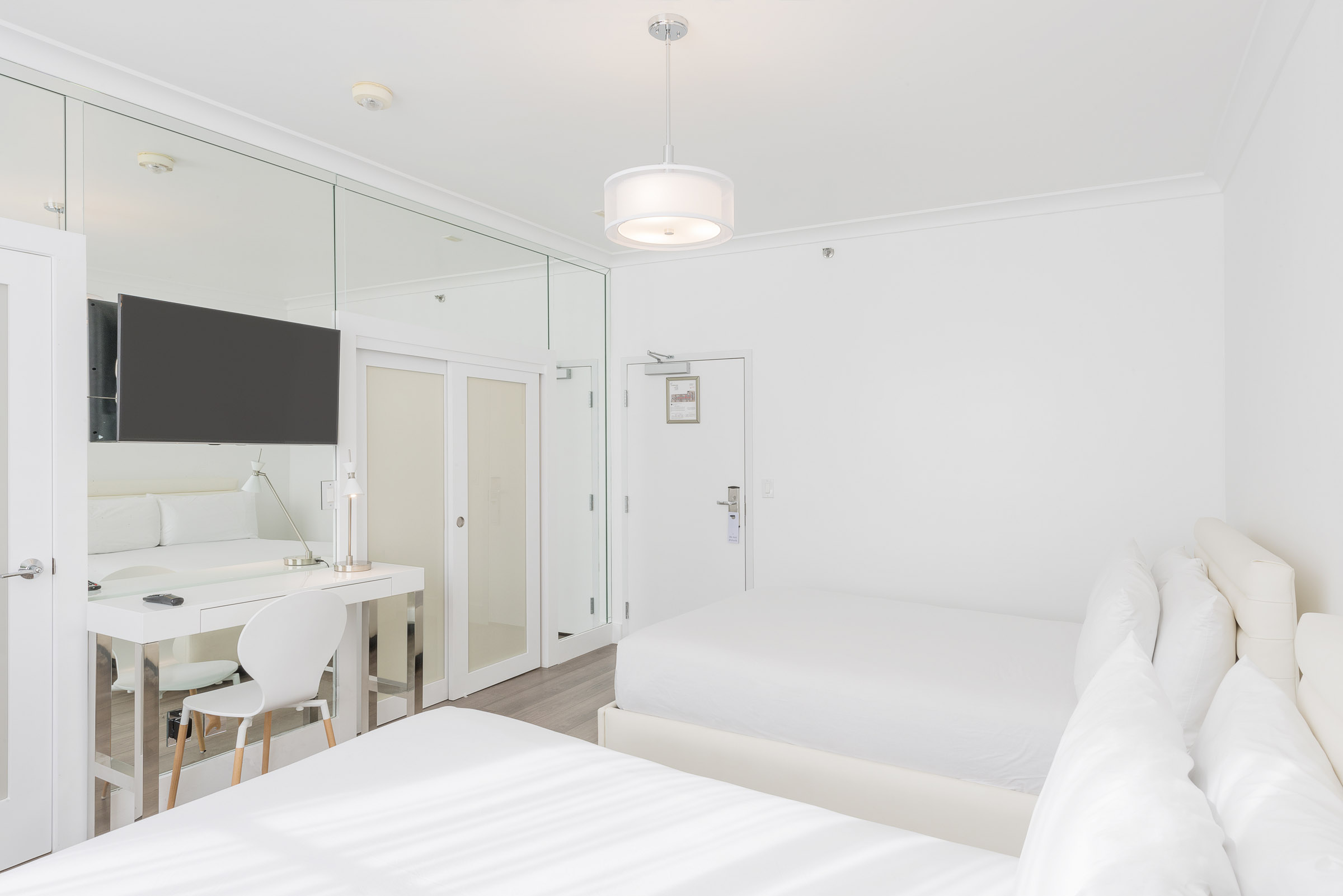 A white room with two beds, a desk, and a TV mounted on a mirrored wall.