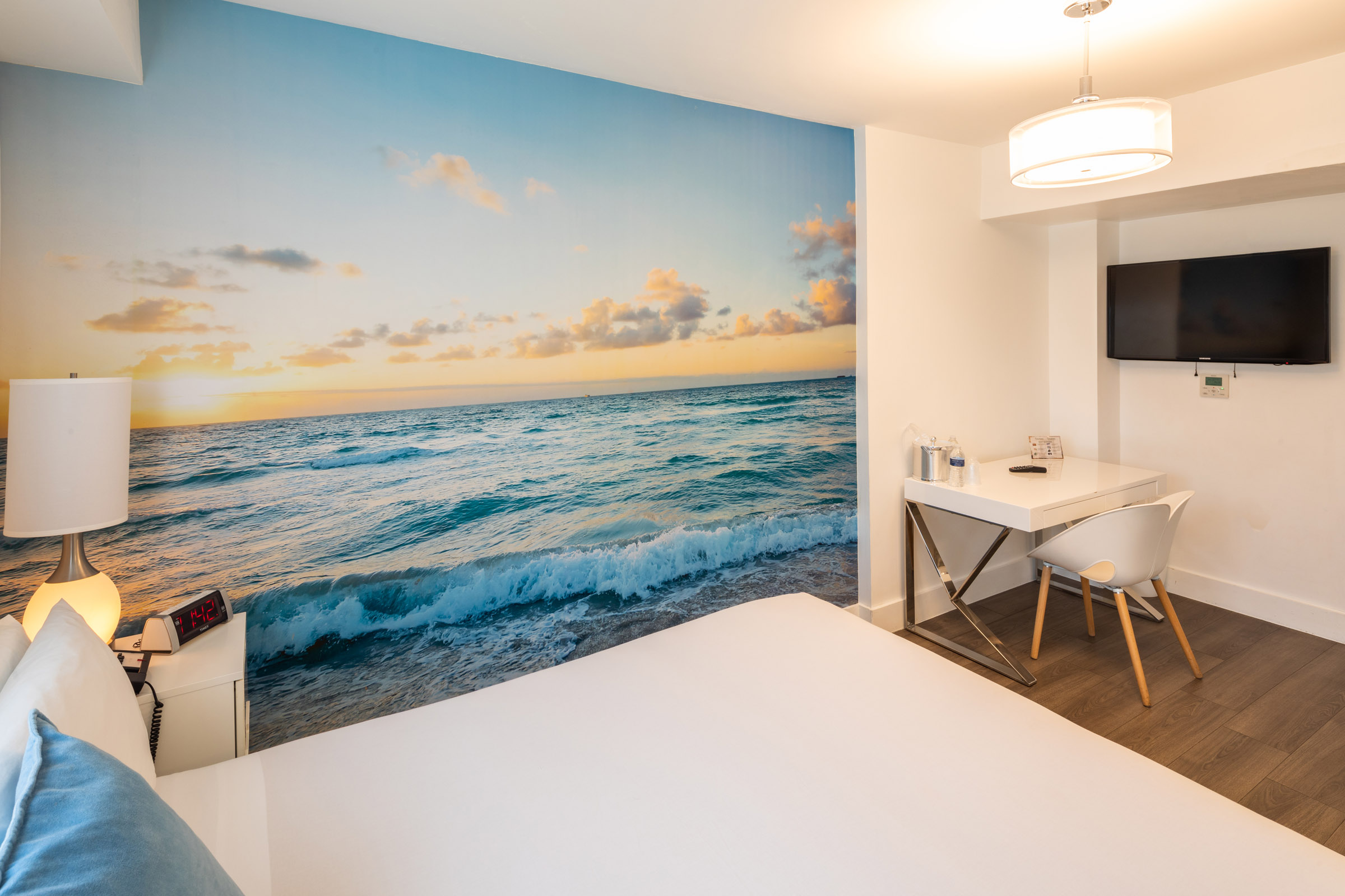 A hotel room with a large wall mural of a beach scene, a bed, desk, chair, and TV.